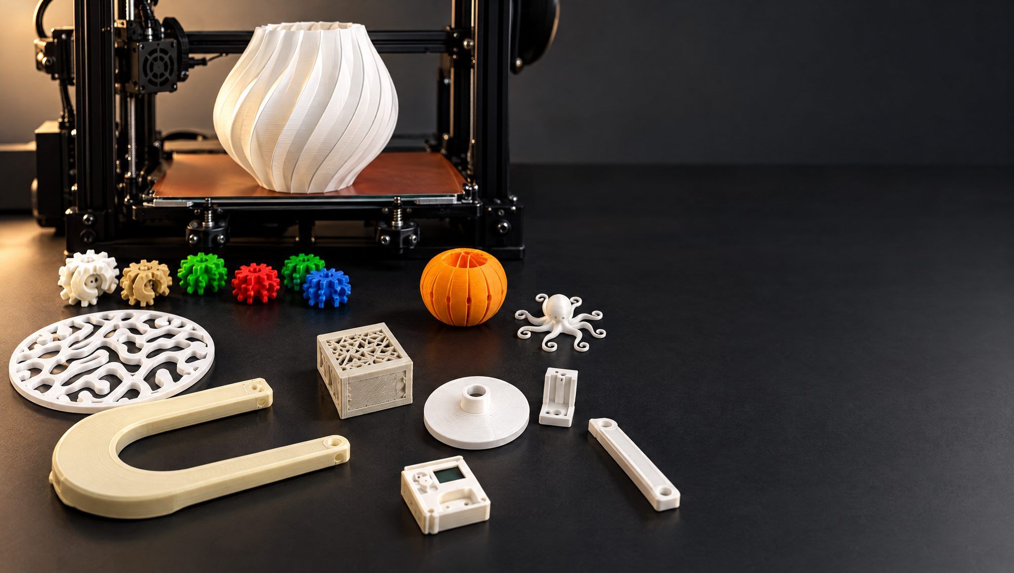 Printed parts and a twisted vase in front of an FDM printer on a dark workbench