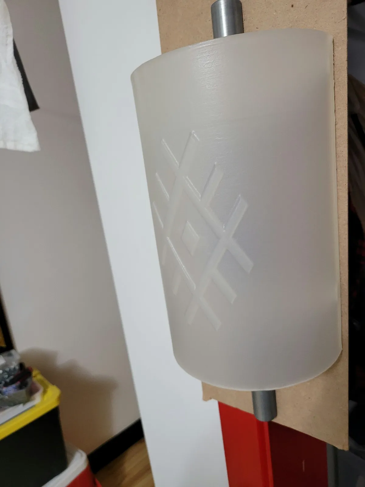 Prototype d’applique éteint, avec un abat-jour blanc courbé, un motif géométrique en losange et un tube métallique vertical.