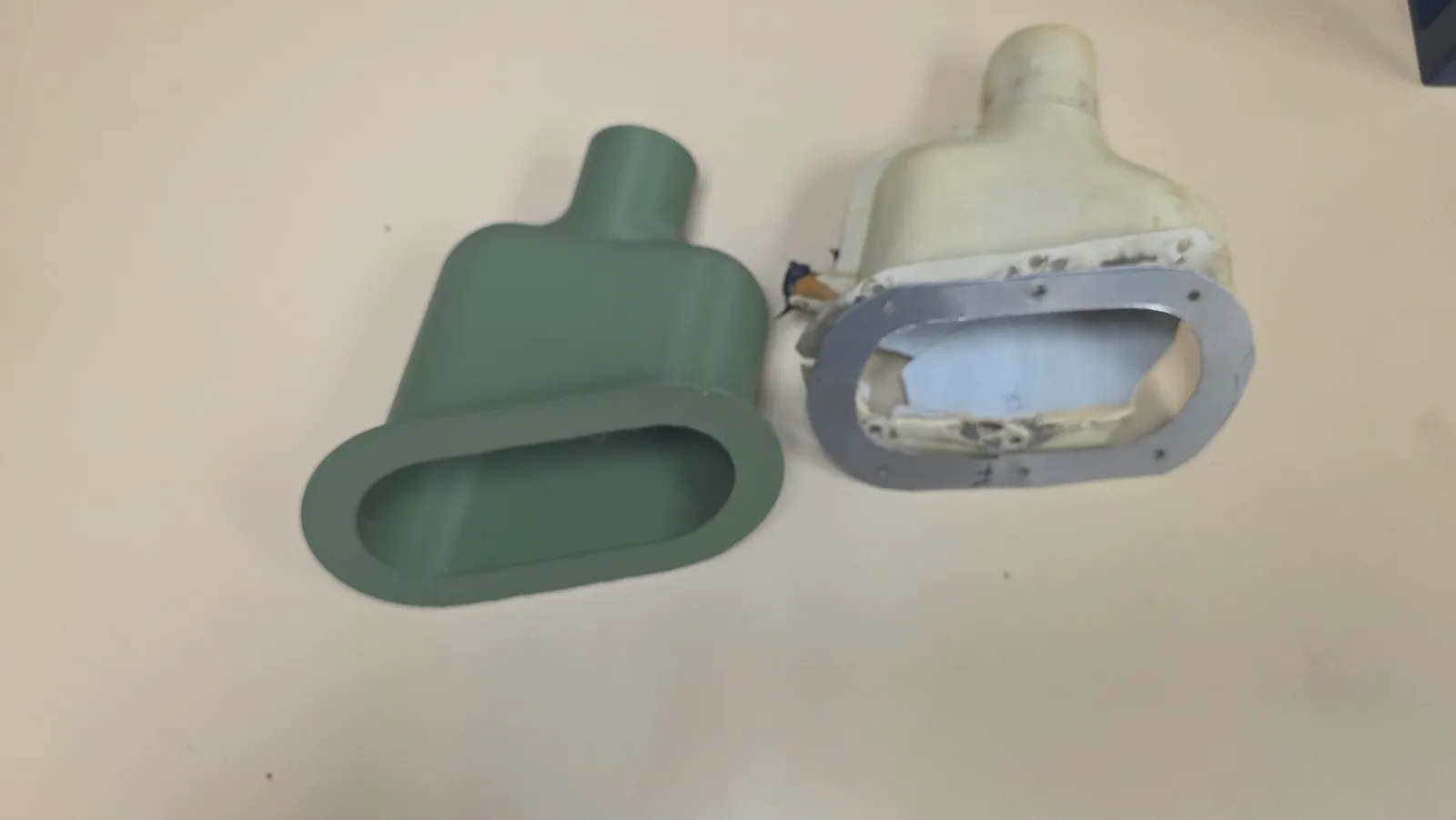 Green 3D-printed replacement duct beside the damaged off-white original, with broad flanged openings and narrower outlets.