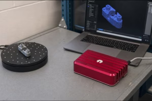 Red aluminium 3D scanner on a lab bench beside a turntable with a printed part and a laptop showing a point cloud